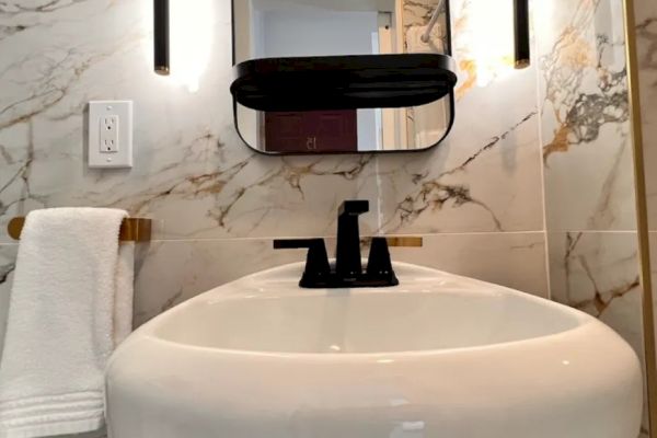 This image shows a modern bathroom with a marble wall, a black faucet, a white sink, a mirror, and wall-mounted lights. A towel hangs on the left.