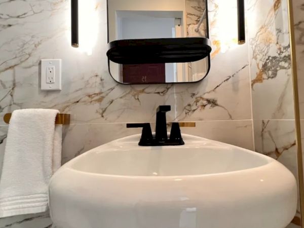 This image shows a modern bathroom with a marble wall, a black faucet, a white sink, a mirror, and wall-mounted lights. A towel hangs on the left.