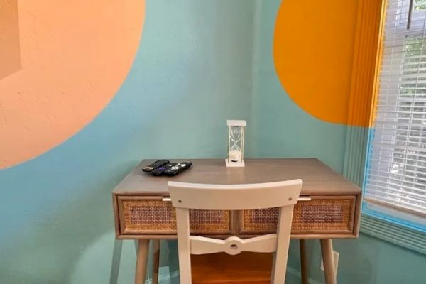 A small desk with a chair, an hourglass, remote control, and bright painted walls with colorful circles, next to a window with blinds.