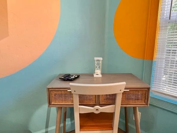 A small desk with a chair, an hourglass, remote control, and bright painted walls with colorful circles, next to a window with blinds.