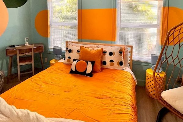 This image shows a colorful bedroom with an orange and light blue wall, featuring a bed, pillows with polka dots, a desk, and a hanging chair.