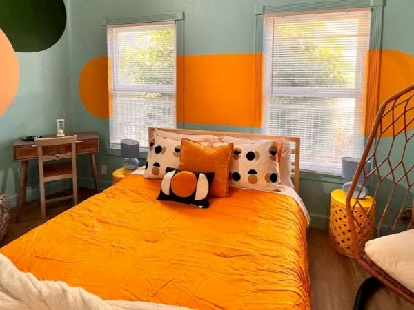 This image shows a colorful bedroom with an orange and light blue wall, featuring a bed, pillows with polka dots, a desk, and a hanging chair.