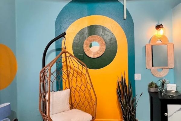 The image shows a colorful room with a hanging chair, geometric wall art, plants, a mirror, and a small black cabinet under a light fixture.