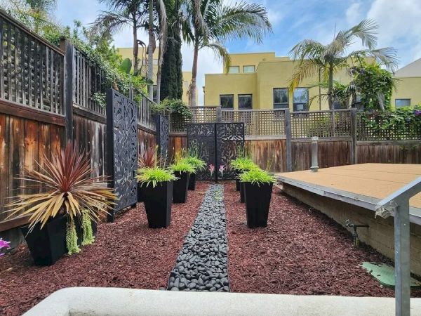 This image shows a landscaped backyard with potted plants, a pebble path, wooden deck, and tall palm trees with a yellow building in the background.