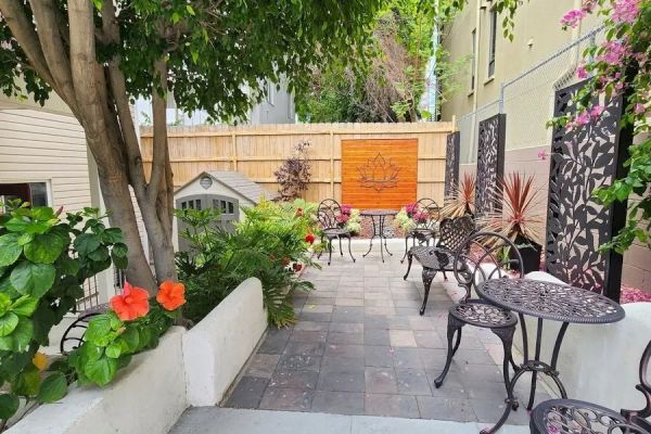 The image shows a small patio with several wrought iron tables and chairs, surrounded by lush greenery and colorful flowers, creating a cozy atmosphere.