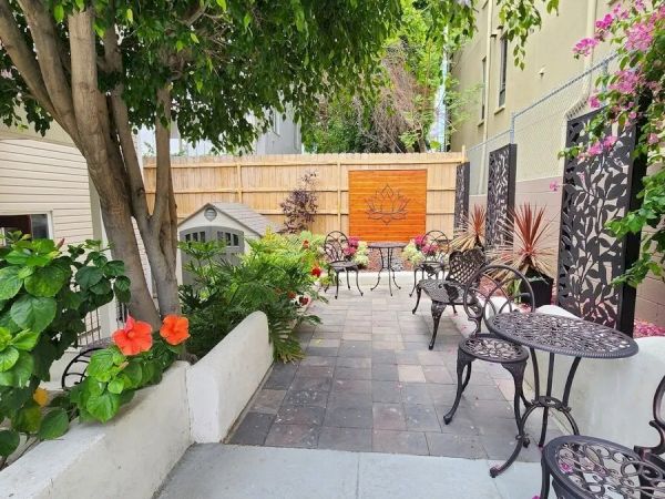 The image shows a small patio with several wrought iron tables and chairs, surrounded by lush greenery and colorful flowers, creating a cozy atmosphere.