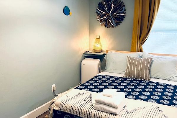 A cozy bedroom with three hanging lights, ocean-themed wall decor, a bed with blue patterned bedding, neatly folded towels, a lamp, and yellow curtains.