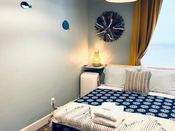 A cozy bedroom with three hanging lights, ocean-themed wall decor, a bed with blue patterned bedding, neatly folded towels, a lamp, and yellow curtains.