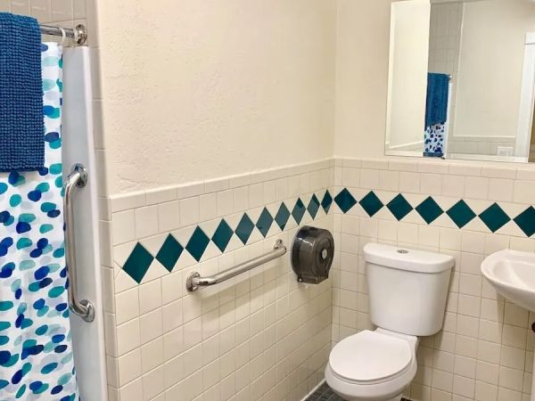 The image shows a bathroom with a toilet, sink, shower with a blue polka dot curtain, grab bar, paper towel dispenser, and a mirror on the wall.
