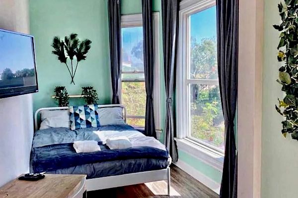 This image shows a cozy bedroom with mint-green walls, a bed with dark blue bedding, a desk, a wall-mounted TV, large windows, and curtains.