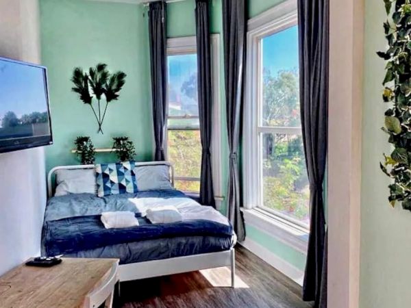 This image shows a cozy bedroom with mint-green walls, a bed with dark blue bedding, a desk, a wall-mounted TV, large windows, and curtains.