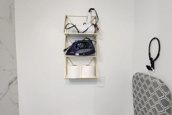 The image shows a bathroom with a wall shelf holding an iron, some cables, and toilet paper. There's an ironing board with a patterned cover.