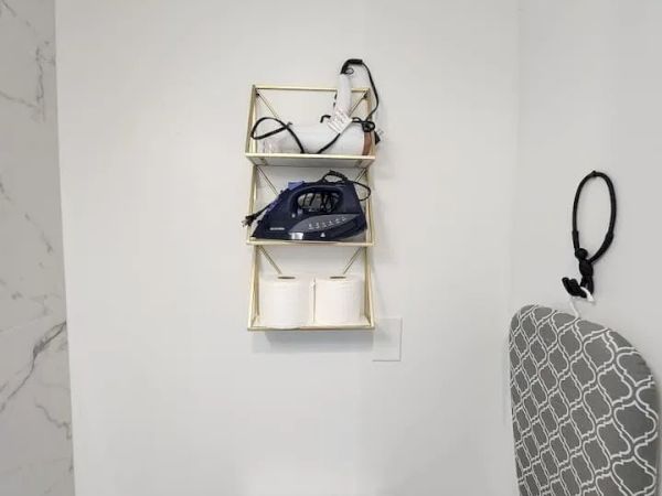 The image shows a bathroom with a wall shelf holding an iron, some cables, and toilet paper. There's an ironing board with a patterned cover.
