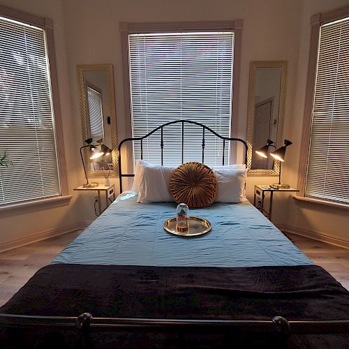 A cozy bedroom features a bed with blue and black linens, flanked by windows with blinds, and bedside tables with lamps and mirrors.