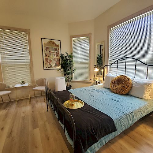 A cozy bedroom with a large bed, pillows, a tray, two chairs, plants, a painting, and a window with blinds. The room has wooden flooring.