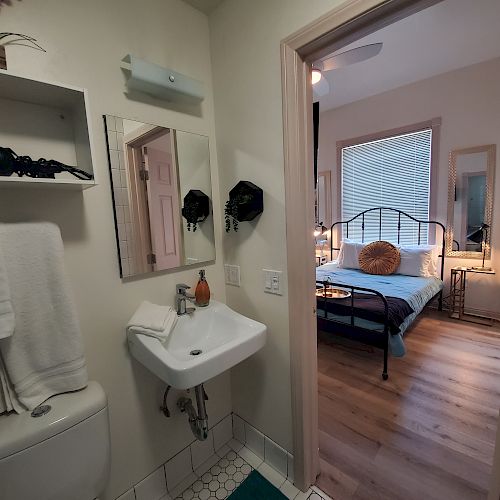 A small bathroom with a sink, mirror, and towels opens to a cozy bedroom with a bed, a large window, and wooden flooring.