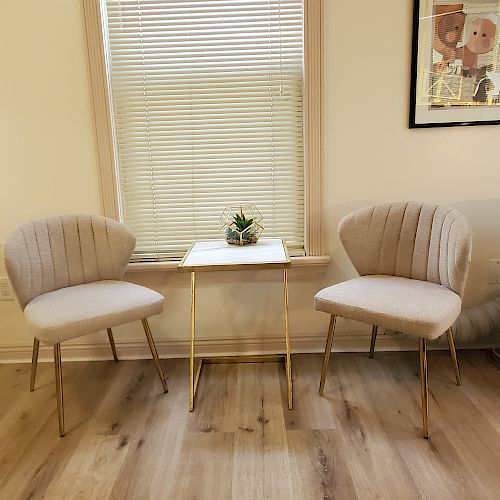 The image shows two beige armchairs and a small side table with a plant, set in front of a window with blinds. A framed picture hangs on the wall.