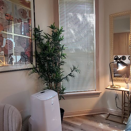 A cozy room with an armchair, a potted plant, a portable air conditioner, a floor lamp next to a full-length mirror, and a large art piece on the wall.