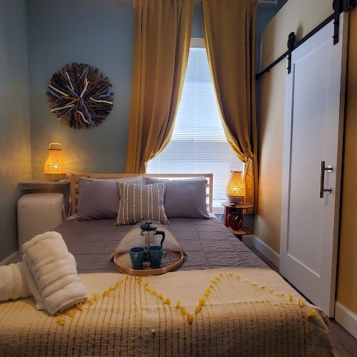 This image shows a cozy bedroom with a bed, pillows, rolled towels, a side table with a lamp, a window with curtains, and a sliding barn door.