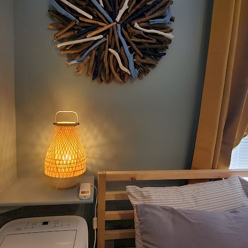 A cozy bedroom corner with a wicker lamp, art piece made of driftwood, blinds, and a bed with striped sheets and multiple pillows ending the sentence.