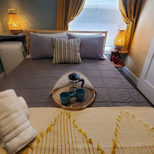 A cozy bedroom features a neatly made bed, folded towels, a tray with a teapot and cups, bedside lamps, and a window with curtains.