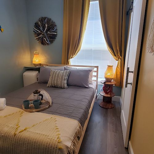 A cozy bedroom with a neatly made bed, decorative pillows, folded towels, wall art, bedside lamps, and a window with curtains.