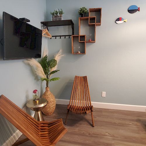 A modern room with wooden chairs, a wall-mounted TV, shelves, artificial plants, a small round table with pampas grass, fish wall art, and towels on a bench.