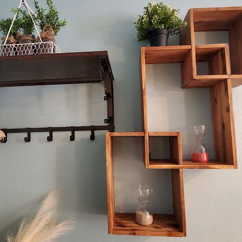 The image shows a minimalist wall setup with hanging wooden shelves, plants, hourglasses, hangers, and a small wall-mounted décor that looks like a red and blue bean.