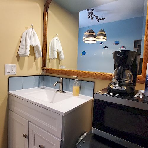 A small kitchenette with a sink, mirror, coffee maker, microwave, coffee creamer, and sugar containers. Towel holders are on the wall.