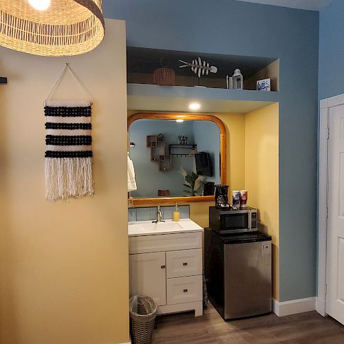 The image shows a small kitchenette area with a sink, a mini-fridge, a microwave, and a mirror. There is a door on the right and a hanging woven decor.