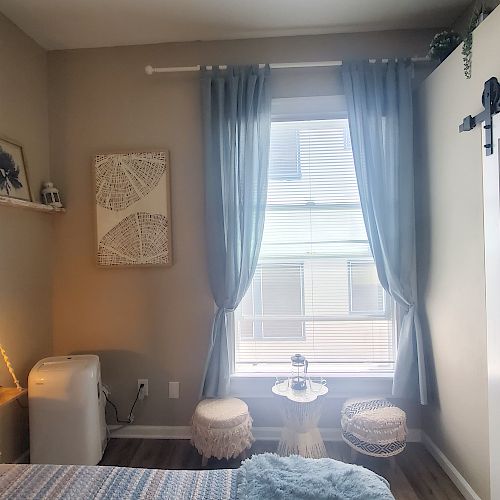 A neatly decorated bedroom with a bed, a hanging light, framed art, a window with curtains, a small table with chairs, a portable AC unit, and a sliding door.