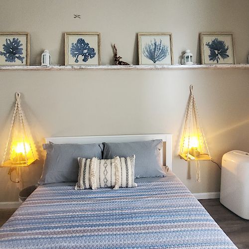 A cozy bedroom with a bed, blue and white bedding, wall art, hanging lights, and a white portable air conditioner on the floor.