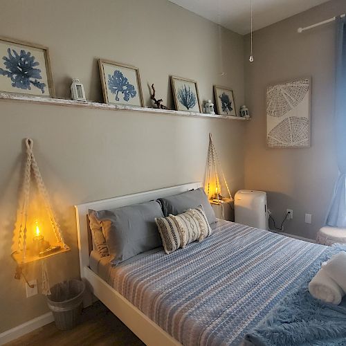 A cozy bedroom with a neatly made bed, blue accents, wall art, soft lighting, and a large window with sheer curtains.