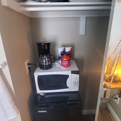 The image shows a small kitchenette area with a microwave, mini fridge, coffee maker, an iron, and some household items on shelves.