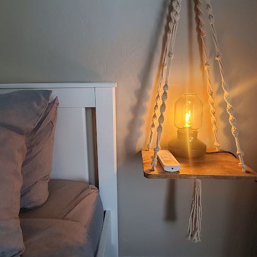 A cozy bedroom scene with a hanging shelf holding a lit lamp and a white remote, next to a bed and a small appliance.