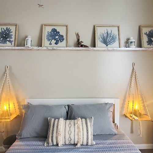 A cozy bedroom with a bed, framed botanical art, two hanging lamps, and decorative items. The room is softly lit and has a serene vibe.