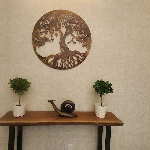 The image shows a wooden console table with two potted plants and a snail figurine, with a metal tree wall art hanging above it.