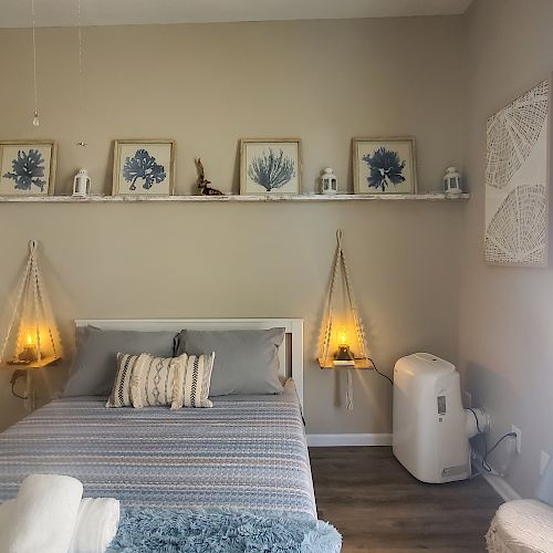 A cozy bedroom with a neatly made bed, wall decor, hanging lights, a small air conditioner, and a window with blue curtains.