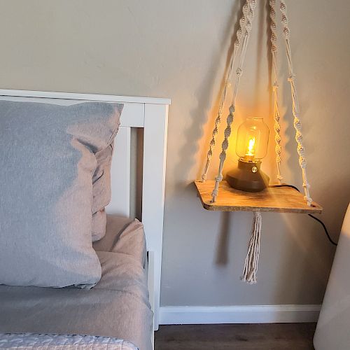 The image shows a bedside with gray bedding, a hanging shelf holding a lamp with a warm light, and a white appliance, possibly an air conditioner.