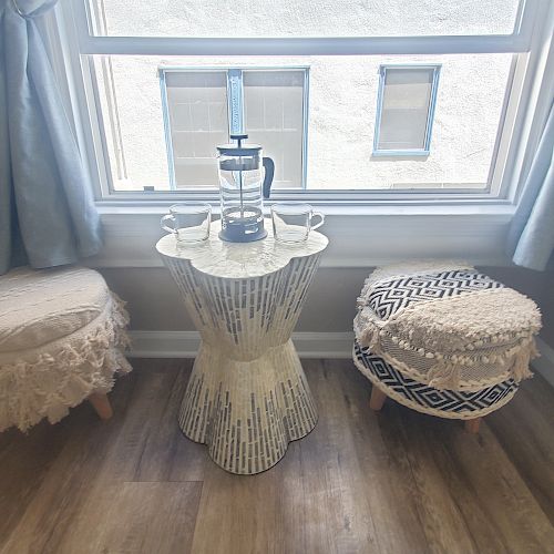 The image shows two cushioned stools and a small decorative table with a teapot, cups, and a French press set, situated by a window with blue curtains.