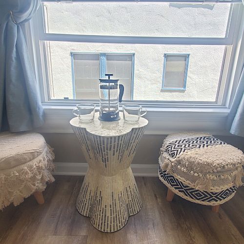 A cozy seating area with two stools, a small table in between, a French press coffee maker, and two cups by a window with light blue curtains.