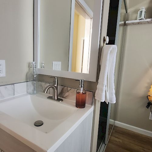 A bathroom with a white sink, towel, mirror, and shelf with candle and decor. The walls are beige, and the floor is wooden.