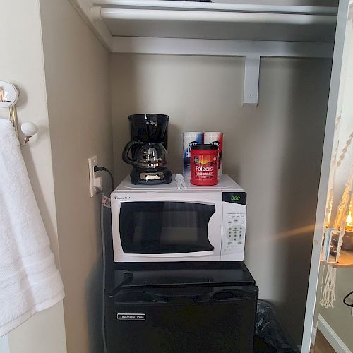 The image shows a small pantry with a mini fridge, microwave, coffee maker, and coffee supplies on a lower shelf; an upper shelf holds miscellaneous items.