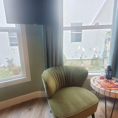 A cozy room with green chair, round table holding a magazine, lantern, and remote, in front of a window with light curtains.