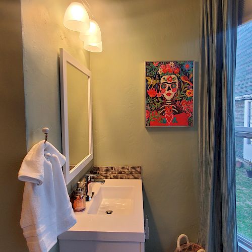 A bathroom with a sink vanity, a mirror with lights, a towel hanging, artwork on the wall, and a window with curtains. A basket is on the floor.