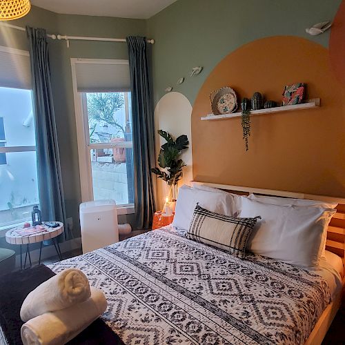 A cozy bedroom with a neatly made bed, patterned bedding, plants, a small round table with chairs near windows, and towels on the bed.