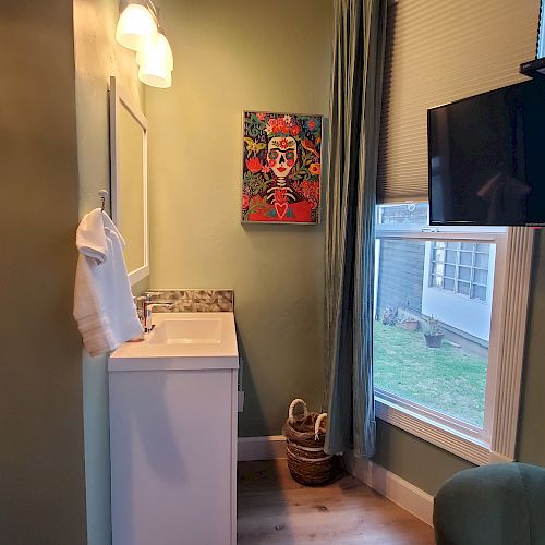 The image shows a small room with a sink, a mirror, a towel, a colorful painting, a wall-mounted TV, a green chair, and a window with curtains.