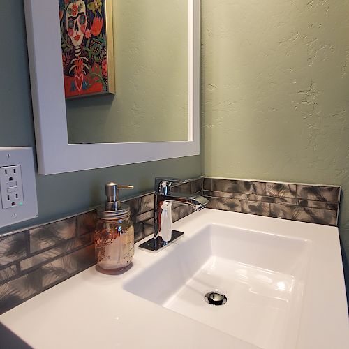 The image shows a small, modern bathroom sink with a mirror above it, a soap dispenser, and decorative wall art.