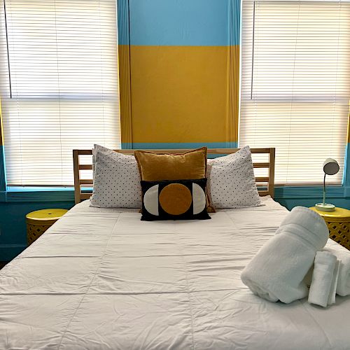 A neatly made bed with decorative pillows and rolled towels. Bright turquoise and yellow wall decor behind, with blinds on both windows ending the sentence.