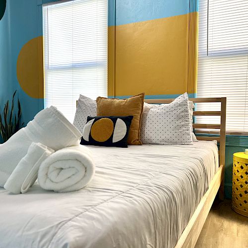 A bedroom with a neatly made bed, white blankets and towels, decorative pillows, a yellow side table with a lamp, and modern wall decor.